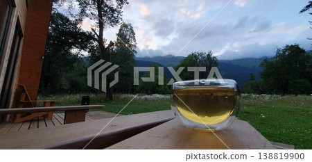 Phone photo of tea in a glass cup on an outdoor terrace with evening views of the Caucasus Mountains. Cozy travel moment showing relaxation, mountain scenery, and quiet atmosphere. Phone photo of tea in a glass cup on an outdoor terrace with evening views of the Caucasus Mountains. Cozy travel moment showing relaxation, mountain scenery, and quiet atmosphere. 138819900
