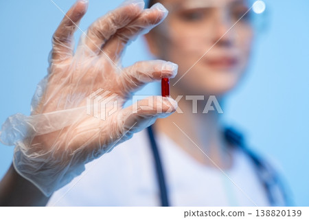 Healthcare professional wearing protective glove holding a small red capsule. Concept for medication, research, and illness prevention Healthcare professional wearing protective glove holding a small red capsule. Concept for medication, research, and illness prevention 138820139