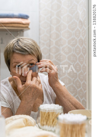 Senior woman applying rejuvenating under-eye patches for a refreshing beauty routine and self-care in a bathroom mirror reflection. Real people, life of an elderly person, daily habits routine Senior woman applying rejuvenating under-eye patches for a refreshing beauty routine and self-care in a bathroom mirror reflection. Real people, life of an elderly person, daily habits routine 138820176