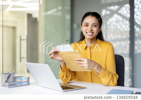 Businesswoman receiving good news from mail, opening a manila envelope with important documents at a desk in a modern office while smiling at the camera Businesswoman receiving good news from mail, opening a manila envelope with important documents at a desk in a modern office while smiling at the camera 138820588