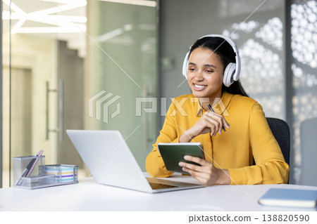 Professional woman wearing headphones and taking notes in a notebook while participating in an online meeting or virtual conference from her office workspace Professional woman wearing headphones and taking notes in a notebook while participating in an online meeting or virtual conference from her office workspace 138820590
