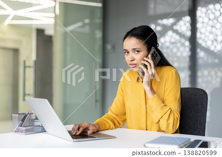 Professional businesswoman sitting at desk in modern home office, typing on laptop and speaking on smartphone while focused on multitasking and virtual communication Professional businesswoman sitting at desk in modern home office, typing on laptop and speaking on smartphone while focused on multitasking and virtual communication 138820599