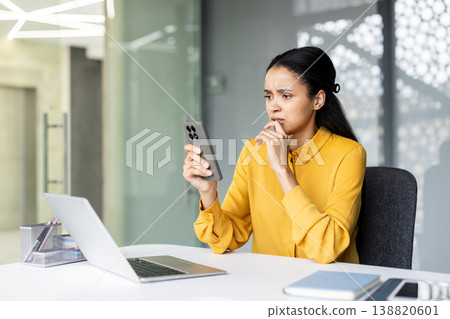 Concerned young businesswoman sitting at her office desk, looking stressed and anxious while reading alarming information or bad news on her mobile phone, with a laptop nearby Concerned young businesswoman sitting at her office desk, looking stressed and anxious while reading alarming information or bad news on her mobile phone, with a laptop nearby 138820601