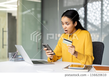 Biracial woman at desk looking shocked at her smartphone while holding a credit card, laptop open, reacting to unexpected online banking charges or potential fraud notification Biracial woman at desk looking shocked at her smartphone while holding a credit card, laptop open, reacting to unexpected online banking charges or potential fraud notification 138820610