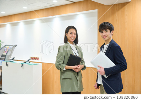Business professionals, both male and female, line up at a company reception desk, smiling and holding laptops and notebooks. 138820860