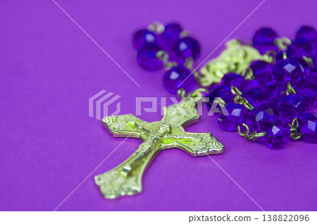 Gold crucifix rosary with purple and light green beads placed on bright violet background in a colorful spiritual object composition. Gold crucifix rosary with purple and light green beads placed on bright violet background in a colorful spiritual object composition. 138822096