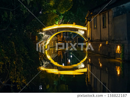 Golden illuminated stone arch bridge casting a vibrant reflection on a quiet canal in a historic Asian water town at night 138822187