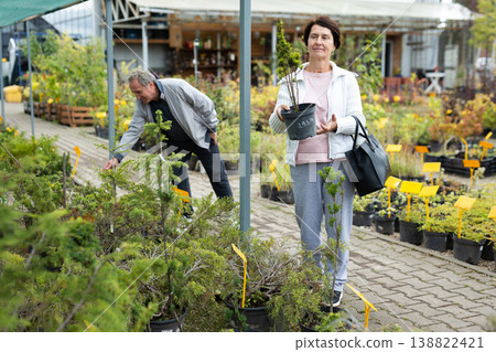 Positive mature couple in casual clothes choosing plants and shrubs in pots while shopping in garden center 138822421