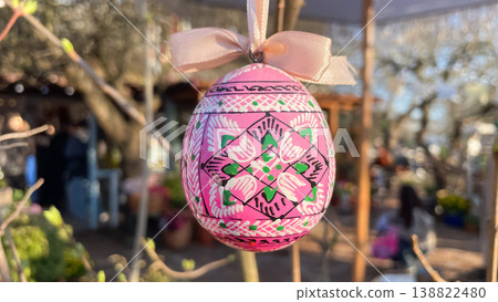Traditional painted Easter egg with white ribbon hanging on budding tree branch in sunny garden Traditional painted Easter egg with white ribbon hanging on budding tree branch in sunny garden 138822480
