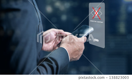 Professional man holding a smartphone with a digital holographic sensitive content warning icon, illustrating data privacy, internet safety, and content moderation policies 138823003