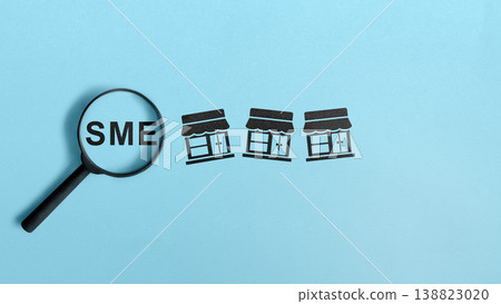 A conceptual image showing a magnifying glass focusing on the acronym SME next to small shop icons on a light blue background, representing business analysis and growth of small businesses 138823020