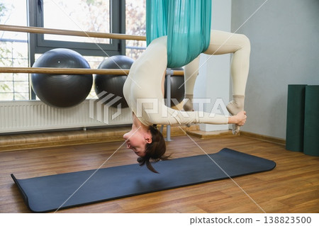 Young woman performing inverted backbend pose using aerial yoga hammock in studio. Young woman performing inverted backbend pose using aerial yoga hammock in studio. 138823500