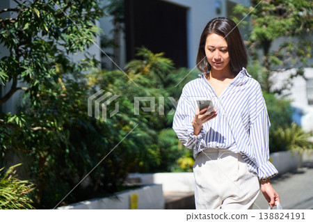 On her way home from shopping, a Japanese woman smiles and enjoys a leisurely stroll on her day off, smartphone in hand. On her way home from shopping, a Japanese woman smiles and enjoys a leisurely stroll on her day off, smartphone in hand. 138824191
