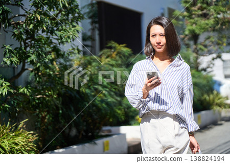On her way home from shopping, a Japanese woman smiles and enjoys a leisurely stroll on her day off, smartphone in hand. On her way home from shopping, a Japanese woman smiles and enjoys a leisurely stroll on her day off, smartphone in hand. 138824194