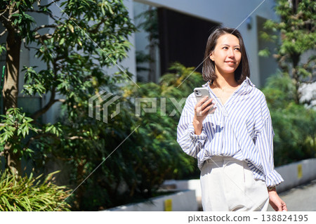 On her way home from shopping, a Japanese woman smiles and enjoys a leisurely stroll on her day off, smartphone in hand. On her way home from shopping, a Japanese woman smiles and enjoys a leisurely stroll on her day off, smartphone in hand. 138824195