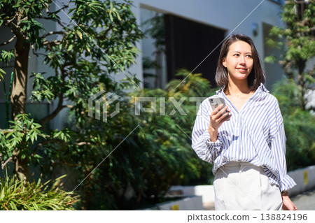 On her way home from shopping, a Japanese woman smiles and enjoys a leisurely stroll on her day off, smartphone in hand. On her way home from shopping, a Japanese woman smiles and enjoys a leisurely stroll on her day off, smartphone in hand. 138824196