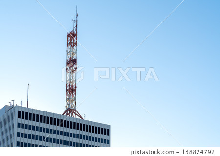 Tall red and white telecommunication tower on top of a modern office building against a clear blue sky. Represents wireless communication, mobile networks, and urban infrastructure. Tall red and white telecommunication tower on top of a modern office building against a clear blue sky. Represents wireless communication, mobile networks, and urban infrastructure. 138824792