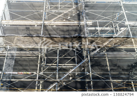 Industrial scaffolding structure with safety netting and metal poles at a construction site. Detailed view of urban building renovation and maintenance equipment in bright daylight. Industrial scaffolding structure with safety netting and metal poles at a construction site. Detailed view of urban building renovation and maintenance equipment in bright daylight. 138824794