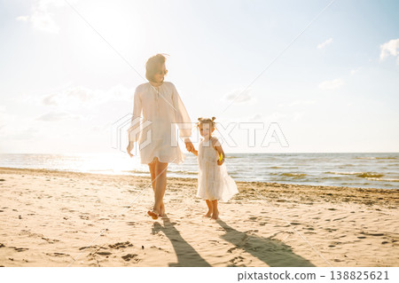 A mother and daughter enjoy a sunny beach stroll, as waves gently lap at their feet during sunset A mother and daughter enjoy a sunny beach stroll, as waves gently lap at their feet during sunset 138825621