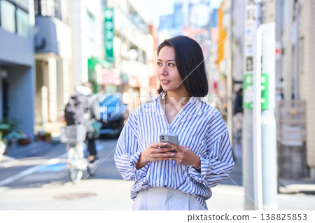 A Japanese woman smiles as she looks at her smartphone while shopping. A stroll through the city on a holiday. A Japanese woman smiles as she looks at her smartphone while shopping. A stroll through the city on a holiday. 138825853