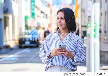 一位日本女子一邊看著手機一邊購物,臉上帶著微笑。這是她在假日裡漫步城市時的照片。 一位日本女子一邊看著手機一邊購物,臉上帶著微笑。這是她在假日裡漫步城市時的照片。 138825854