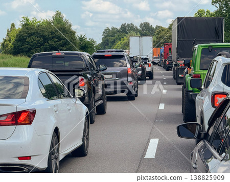 A long line of cars and delivery trucks moves slowly along a highway during heavy traffic. Brake lights glow as vehicles queue between lanes surrounded by greenery. A long line of cars and delivery trucks moves slowly along a highway during heavy traffic. Brake lights glow as vehicles queue between lanes surrounded by greenery. 138825909