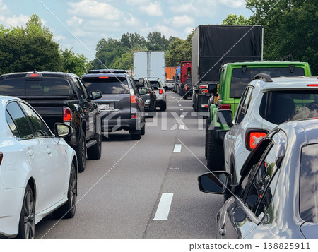 Cars and trucks fill multiple lanes of a busy highway creating heavy traffic congestion. Vehicles move slowly under a cloudy sky surrounded by green trees. 138825911
