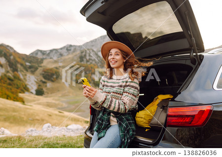 Young traveler woman sits on the rear of her car, smiling and using phone in scenic mountainous area Young traveler woman sits on the rear of her car, smiling and using phone in scenic mountainous area 138826036