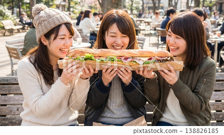 三位日本女性面帶微笑地坐在公園長椅上，分享著一個巨大的法國長棍三明治。 138826185