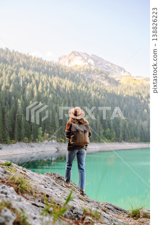 Back view young woman with backpack looking the view of the mountain lake in sunny weather. Back view young woman with backpack looking the view of the mountain lake in sunny weather. 138826223