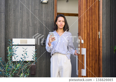 A Japanese woman leaves the front door of a detached house, smartphone in hand. A Japanese woman leaves the front door of a detached house, smartphone in hand. 138826665