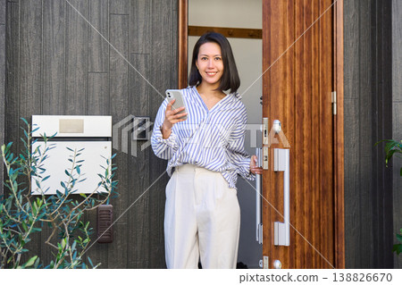 A Japanese woman leaves the front door of a detached house, smartphone in hand. A Japanese woman leaves the front door of a detached house, smartphone in hand. 138826670