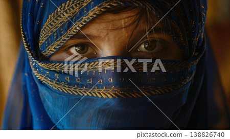 A woman wearing a vibrant blue hijab adorned with intricate gold patterns gazes intensely 138826740