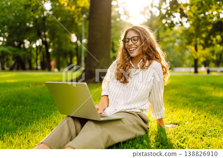 Happy Woman with laptop sit green grass at park. Studying outdoors. Freelancing concept Happy Woman with laptop sit green grass at park. Studying outdoors. Freelancing concept 138826910