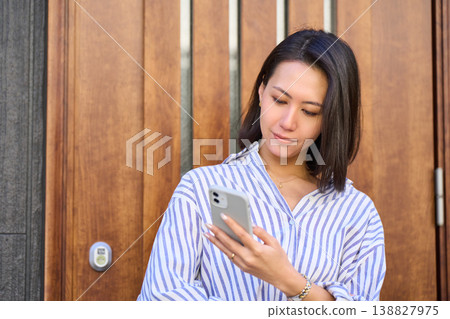 A Japanese woman smiles as she operates her smartphone in front of the entrance of a detached house before going out. A Japanese woman smiles as she operates her smartphone in front of the entrance of a detached house before going out. 138827975
