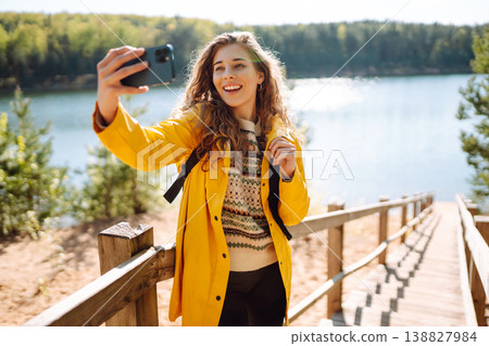 Happy woman with phone in hands taking selfie in forest. Young tourist enjoying spring day near lake 138827984