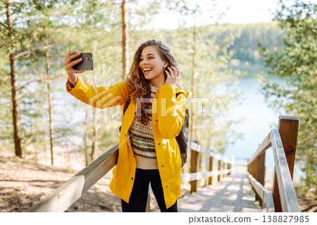 Happy woman with phone in hands taking selfie in forest. Young tourist enjoying spring day near lake 138827985