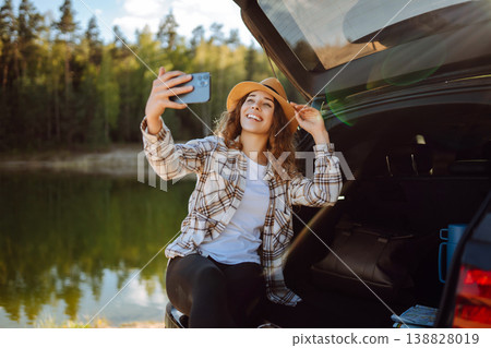 Selfie in nature. Happy woman sits in trunk of car, uses smartphone. Concept of traveling, blogging. Selfie in nature. Happy woman sits in trunk of car, uses smartphone. Concept of traveling, blogging. 138828019