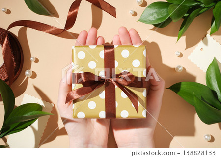 Hands holding a polka dot gift box with a brown ribbon over a beige background with leaves and pearls. 138828133
