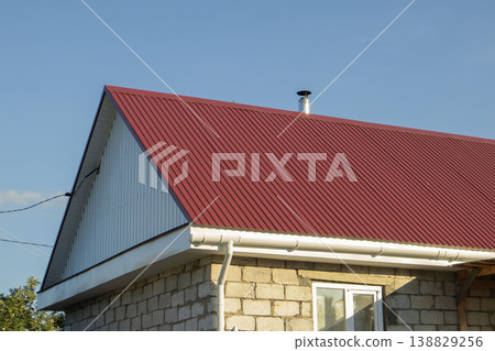 Red metal roof with vertical ribbing on a stone house under clear blue sky, chimney visible on the roofline, showcasing modern architectural design elements Red metal roof with vertical ribbing on a stone house under clear blue sky, chimney visible on the roofline, showcasing modern architectural design elements 138829256