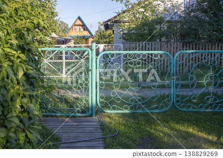 Decorative turquoise metal gate with intricate patterns leading to a garden path, surrounded by greenery and a wooden walkway in a residential area Decorative turquoise metal gate with intricate patterns leading to a garden path, surrounded by greenery and a wooden walkway in a residential area 138829269