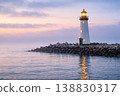 Lighthouse on rocky shore at sunset with glowing light over calm sea 138830317