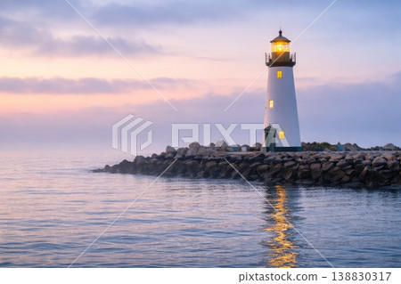 Lighthouse on rocky shore at sunset with glowing light over calm sea Lighthouse on rocky shore at sunset with glowing light over calm sea 138830317