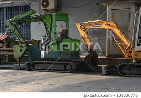 A verdant green excavator sits in stationary repose, its open cab and folded arm capturing the functional grit of the site. To its right, a burnished gold Komatsu arm enters the frame, catching the A verdant green excavator sits in stationary repose, its open cab and folded arm capturing the functional grit of the site. To its right, a burnished gold Komatsu arm enters the frame, catching the 138830778