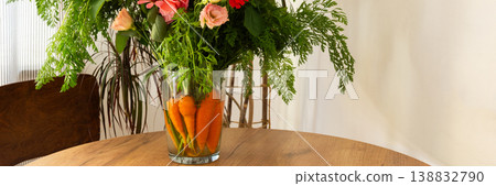 A bouquet features roses, gladioli, zinnias, and eustomas. Carrots with green tops sit in water, placed on a wooden table in a bright room, banner A bouquet features roses, gladioli, zinnias, and eustomas. Carrots with green tops sit in water, placed on a wooden table in a bright room, banner 138832790