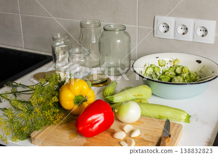 Cucumbers, peppers, and zucchini are being prepared and placed in jars for canning. Fresh ingredients are cut and organized on a clean kitchen counter for preserving 138832823