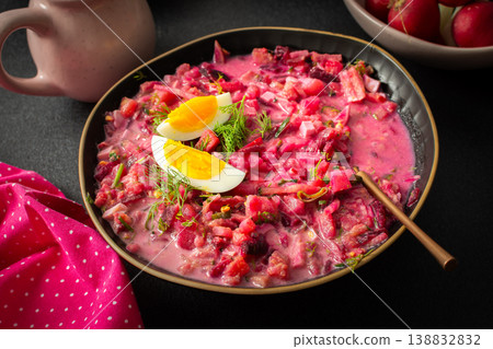 A colorful bowl of beet soup features chopped vegetables, sliced eggs, and fresh herbs. The setting includes a jug of cream, a lemon, and radishes, creating a cozy atmosphere A colorful bowl of beet soup features chopped vegetables, sliced eggs, and fresh herbs. The setting includes a jug of cream, a lemon, and radishes, creating a cozy atmosphere 138832832