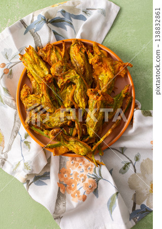 Golden-brown deep-fried zucchini flowers are arranged on a vibrant plate. The dish is set on a floral-patterned napkin, enhancing its appetizing appearance 138832861