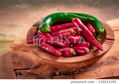 Fresh green and red peppers arranged in a wooden bowl on a rustic wooden board placed on a textured surface Fresh green and red peppers arranged in a wooden bowl on a rustic wooden board placed on a textured surface 138833290