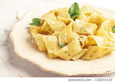 Fresh papardelle pasta, with basil and pesto sauce, is served, on a white plate, with a fork held by hand 138833329